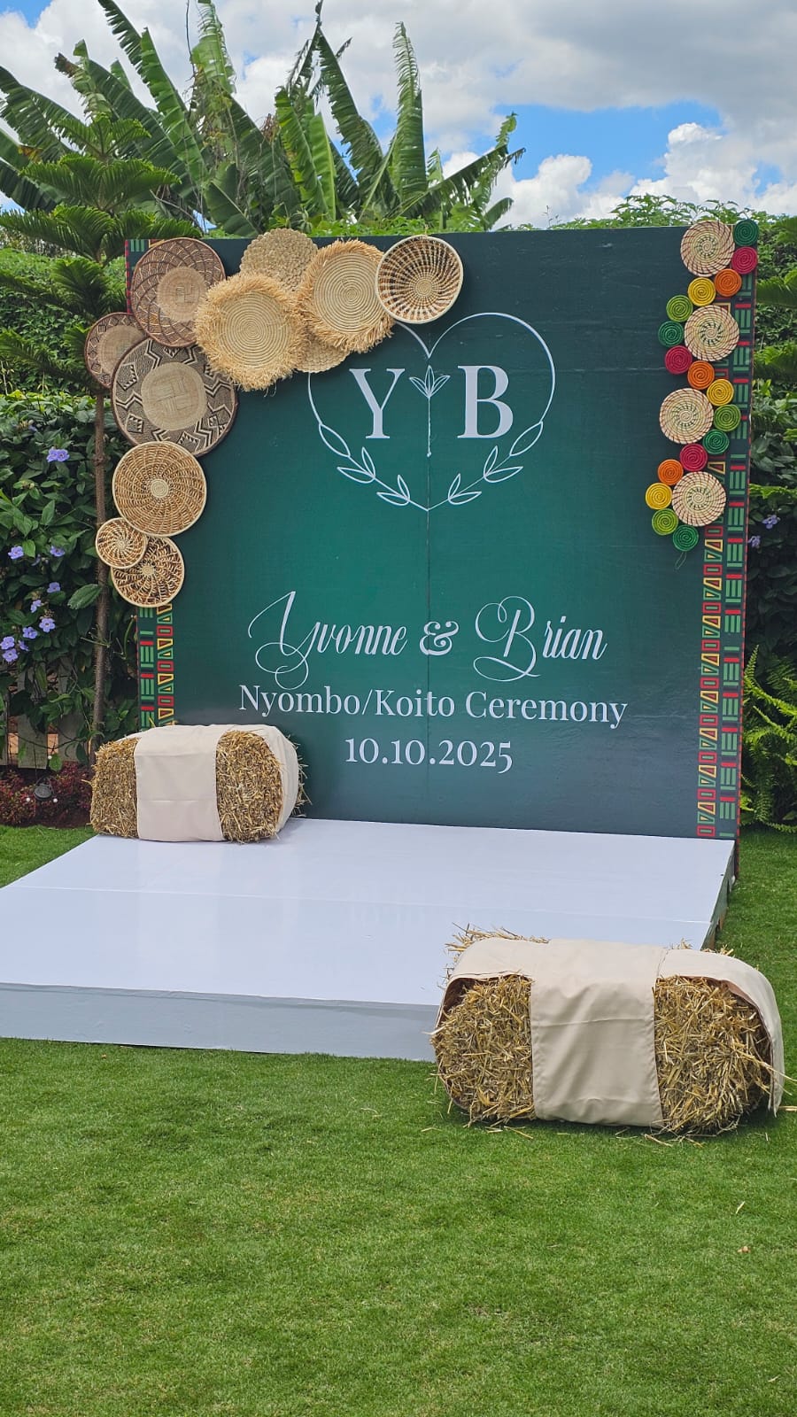 Green traditional ceremony backdrop with woven baskets and hay bale seating on the garden lawn