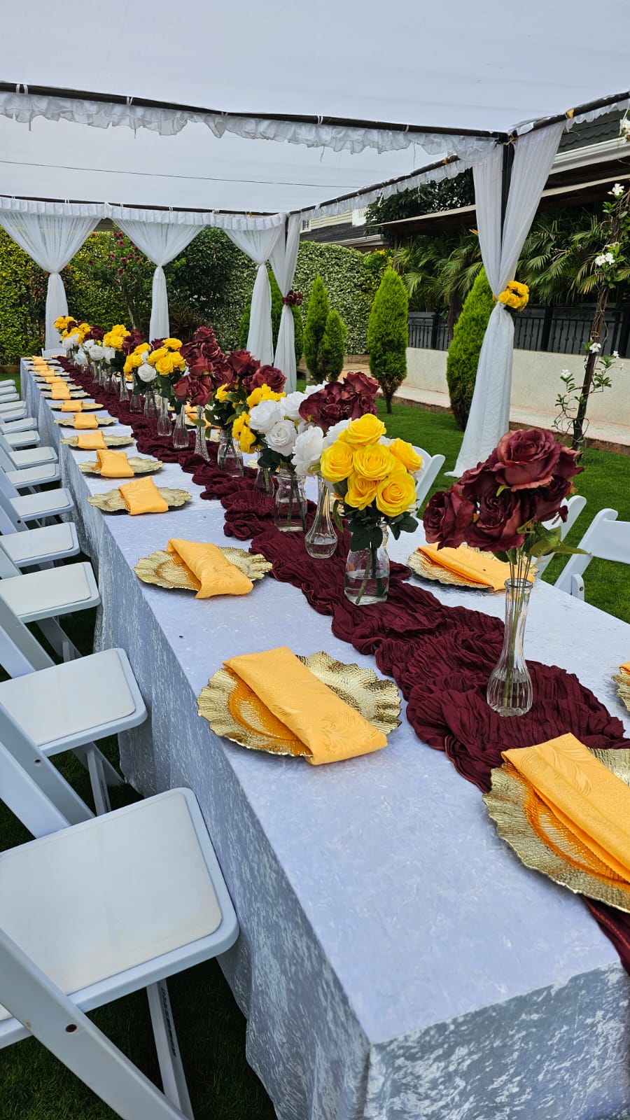 Long garden table dressed in white with burgundy runners, gold chargers and rose arrangements under a draped canopy